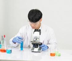 asiático masculino estudiante científico vistiendo un del doctor vestido en el laboratorio mirando mano a químico. causado por mezcla reactivos en científico investigación laboratorios con prueba tubos y microscopio en el mesa foto