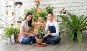Gardener young Asian man woman two person sit floor and smiling looking hand holding help decorate the tree leaf green in calm work shop home plant white wall. hobby job happy and care concept photo