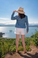 posterior ver de mujer en Paja sombrero, mezclilla chaqueta, blanco pantalones cortos y sandalias soportes en antecedentes Oceano foto