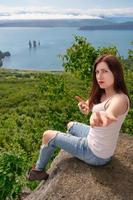 Woman sits on rocky shore in mountains against panoramic view of green forest, ocean on summer day photo