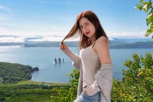 Adult woman in light T-shirt standing on mountain shore against panoramic view of sea on summer day photo