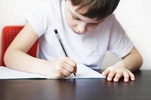 el chico se sienta a el mesa y escribe en un cuaderno. niño se sienta y lo hace deberes en un blanco antecedentes foto