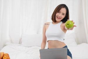 Portrait of a white Asian woman with tattoos sitting holding green apples and fruits in a healthy lifestyle. Relaxing in bed and playing on my laptop for the weekend. holiday concept. photo