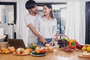 Young Asian couple cooking with fruits and vegetables and using laptop in the kitchen To cook food together within the family happily, family concept. photo