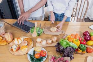 Young Asian couple cooking with fruits and vegetables and using laptop in the kitchen To cook food together within the family happily, family concept. photo