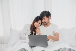 Young happy asian couple using laptop in bed watching movie, romantic time to enhance family relationship, family concept. photo