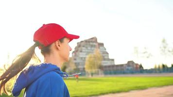 donna con senza fili cuffie e smartphone sceglie musica e corre attraverso il stadio a tramonto. lento movimento video