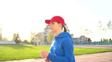 mujer con inalámbrico auriculares y teléfono inteligente elige música y carreras mediante el estadio a puesta de sol. lento movimiento video