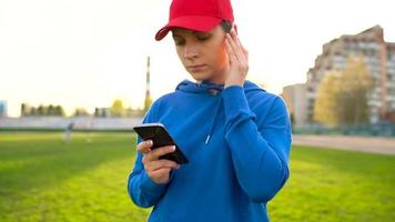 mujer con inalámbrico auriculares y teléfono inteligente elige música y carreras mediante el estadio a puesta de sol. lento movimiento video