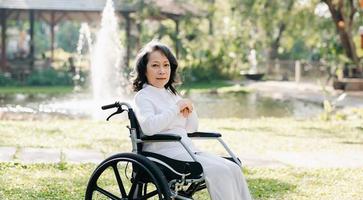 Happy elderly woman sitting on wheelchair outdoor in park relax your mind with green nature. Nursing home hospital garden concept. photo