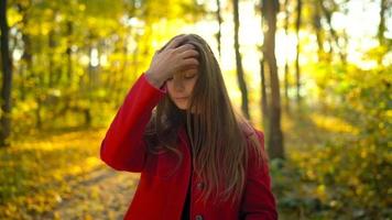 Portrait of a beautiful pensive girl in a red coat with a yellow maple leaf in the background in the autumn forest. Slow motion video