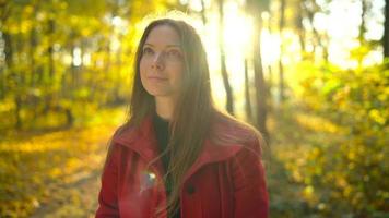 Portrait of a beautiful pensive girl in a red coat with a yellow maple leaf in the background in the autumn forest. Slow motion video