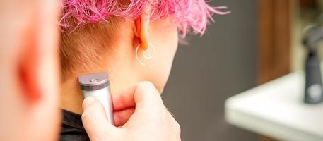 Back view of hairdresser's hand shaving nape and neck with electric trimmer of young caucasian woman with short pink hair in beauty salon. photo