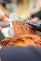 Closeup of master's hand with blow-drying and hairbrush blowing female red hair in a salon. photo