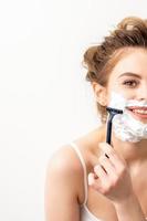 Portrait of beautiful young smiling caucasian woman shaves face with razor on white background. photo