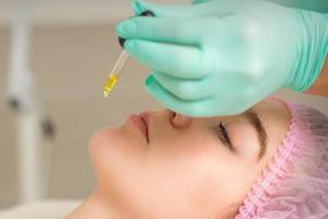Close up of beautician holding pipette and applying oil serum drop on the face of a young woman in spa salon. photo