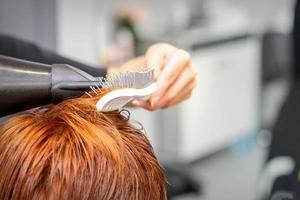 Closeup of master's hand with blow-drying and hairbrush blowing female red hair in a salon. photo