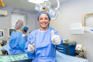 Portrait of female woman nurse surgeon OR staff member dressed in surgical scrubs gown mask and hair net in hospital operating room theater making eye contact smiling thumbs up photo