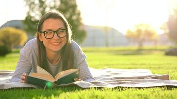 Girl in glasses reading book lying down on a blanket in the park at sunset video