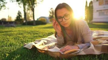 menina dentro óculos lendo livro deitado baixa em uma cobertor dentro a parque às pôr do sol video