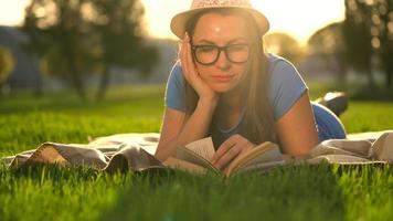 fille dans des lunettes en train de lire livre mensonge vers le bas sur une couverture dans le parc à le coucher du soleil video