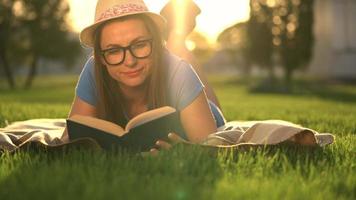 fille dans des lunettes en train de lire livre mensonge vers le bas sur une couverture dans le parc à le coucher du soleil video