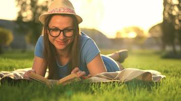 fille dans des lunettes en train de lire livre mensonge vers le bas sur une couverture dans le parc à le coucher du soleil video