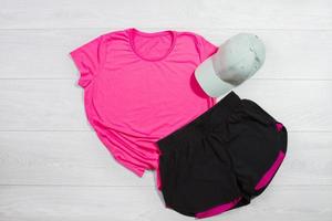 Pink female sport run fitness t shirt mockup flat lay on wooden background. Summer pilates stratching saccessories. Sporty trousers, hat. Top front view t-shirt.T-shirt Template blank copy space photo