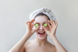 Beauty portrait of woman in towel on head with white nourishing mask or creme on face holding cucumber slices, white background isolated photo