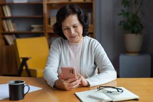 European middle aged senior woman holding using smartphone touch screen typing scroll page. Stylish older mature 60s lady with cell phone using internet social media apps at home. Shopping online. photo