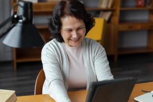 Happy middle aged senior woman holding tablet talk on video call with friends family. Laughing mature old senior grandmother having fun talking speaking with grown up children online. photo