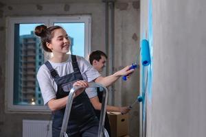 Couple in new home during repair works painting wall together. Happy family holding paint roller painting wall with blue color paint in new house. Home renovation DIY renew home concept. photo