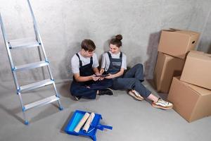 casa bricolaje renovación concepto. joven contento Pareja sentado en piso mirando hogar reparar plan en tableta adentro. mujer demostración a hombre plan interior diseño en línea. Pareja en nuevo hogar durante reparar obras. foto