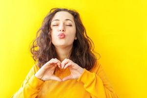 Love, heart shape, peace. Beauty portrait young happy positive woman showing heart sign with hands on yellow background isolated. European girl. Positive human emotion facial expression body language. photo