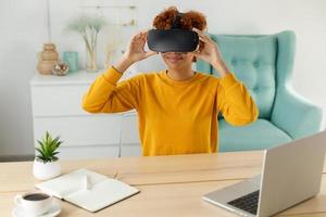 African american young woman wearing using virtual reality metaverse VR glasses headset at home. Girl puts on virtual reality helmet. Technology simulation hi-tech videogame concept. photo