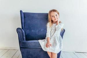 Sweet little girl in white dress sitting on modern cozy blue chair relaxing in white bright living room at home indoors. Childhood schoolchildren youth relax concept. photo