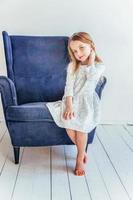 Sweet little girl in white dress sitting on modern cozy blue chair relaxing in white bright living room at home indoors. Childhood schoolchildren youth relax concept. photo