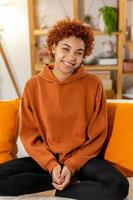 hermosa chica afroamericana con peinado afro sonriendo sentada en un sofá en casa interior. joven africana con cabello rizado riendo. libertad felicidad despreocupada gente feliz concepto. foto