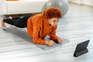 aptitud rutina de ejercicio capacitación. joven sano ajuste africano niña haciendo tablón ejercicio en yoga estera en piso a hogar. atlético mujer en ropa de deporte formación pilates deporte y aptitud física. foto