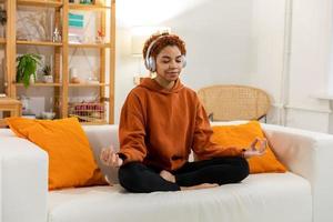 Yoga mindfulness meditation. Young healthy african girl practicing yoga at home. Woman sitting in lotus pose on yoga mat meditating smiling relaxing indoor. Girl doing breathing practice, yoga at home photo