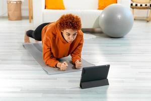 aptitud rutina de ejercicio capacitación. joven sano ajuste africano niña haciendo tablón ejercicio en yoga estera en piso a hogar. atlético mujer en ropa de deporte formación pilates deporte y aptitud física. foto