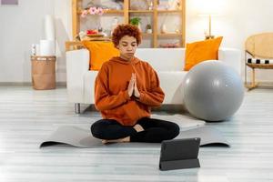 Yoga mindfulness meditation. Young healthy african girl practicing yoga at home. Woman sitting in lotus pose on yoga mat eyes close hands in prayer meditating indoor. Girl doing breathing practice. photo