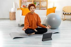 yoga atención plena meditación. joven sano africano niña practicando yoga a hogar. mujer sentado en loto actitud en yoga estera meditando sonriente relajante interior. niña haciendo respiración práctica, yoga a hogar foto