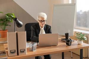 Confident stylish european middle aged senior woman using laptop at workplace. Stylish older mature 60s gray haired lady businesswoman sitting at office table. Boss leader teacher professional worker. photo