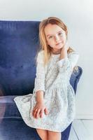 Sweet little girl in white dress sitting on modern cozy blue chair relaxing in white bright living room at home indoors. Childhood schoolchildren youth relax concept. photo