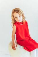 Sweet little girl in red dress sitting on chair against white wall at home, relaxing in white bright living room indoors. Childhood schoolchildren youth relax concept. photo