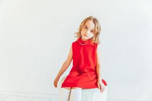 Sweet little girl in red dress sitting on chair against white wall at home, relaxing in white bright living room indoors. Childhood schoolchildren youth relax concept. photo