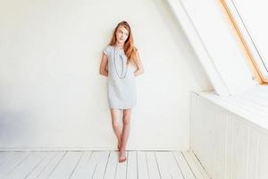 Happy teenage girl smiling. Portrait young happy positive teen woman in grey dress at home in bright room against white wall. European woman. Positive human emotion facial expression body language. photo