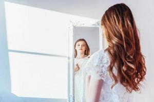 Beauty makeup morning rutine love yourself concept. Young teenage girl looking at reflection in mirror. Young positive woman wearing white dress posing in bright light room against white wall. photo