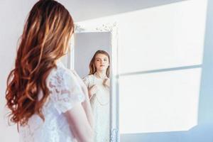 Beauty makeup morning rutine love yourself concept. Young teenage girl looking at reflection in mirror. Young positive woman wearing white dress posing in bright light room against white wall. photo